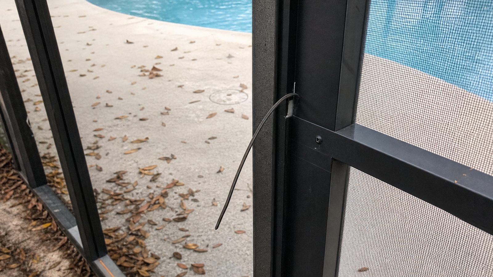 Close view of screen enclosure door frame with loose spline and leaf debris on pool deck